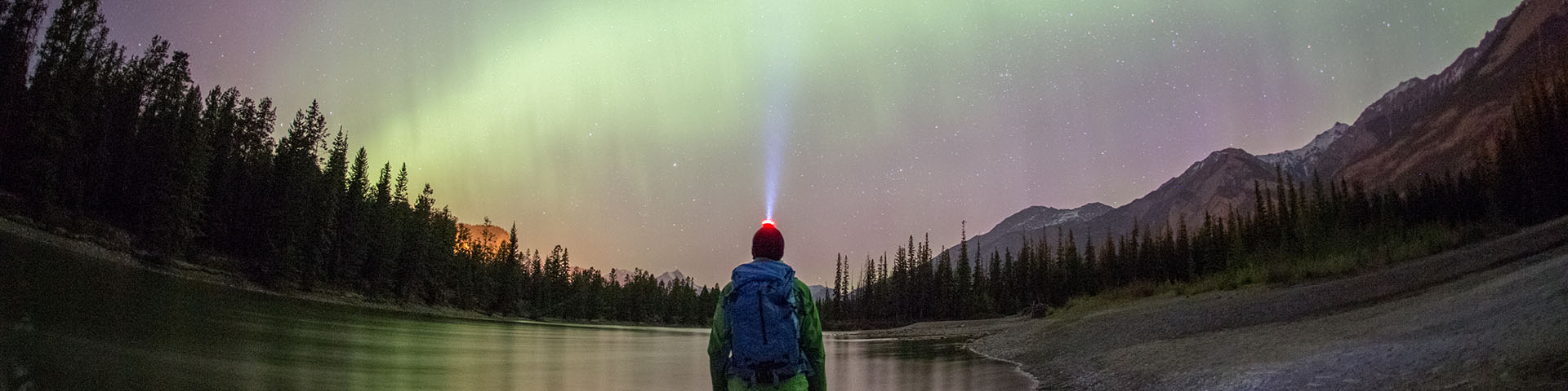 An adult gazes at the Northern Lights on the edge of a lake with mountains and forest in the background at Jasper National Park.
