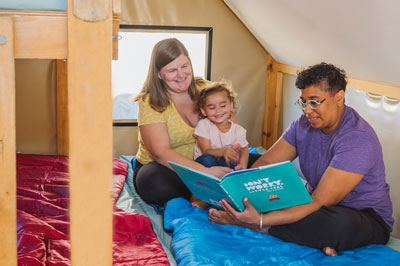 Des parents et leur enfant lisent un livre. EIles se trouvent dans une oTENTik, assis sur un lit superposé recouvert de sacs de couchage.