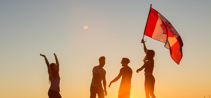 Four young adults celebrate Canada Day by raising a Canadian flag to the sky as the sun sets.