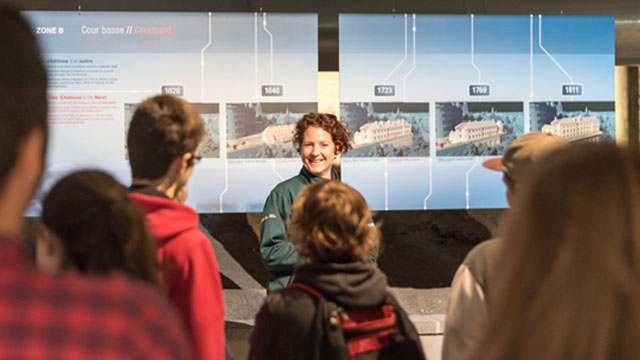 Une guide de Parcs Canada devant un tableau au lieu historique national de Forts-et-Châteaux-Saint-Louis donne des explications à des visiteurs. 