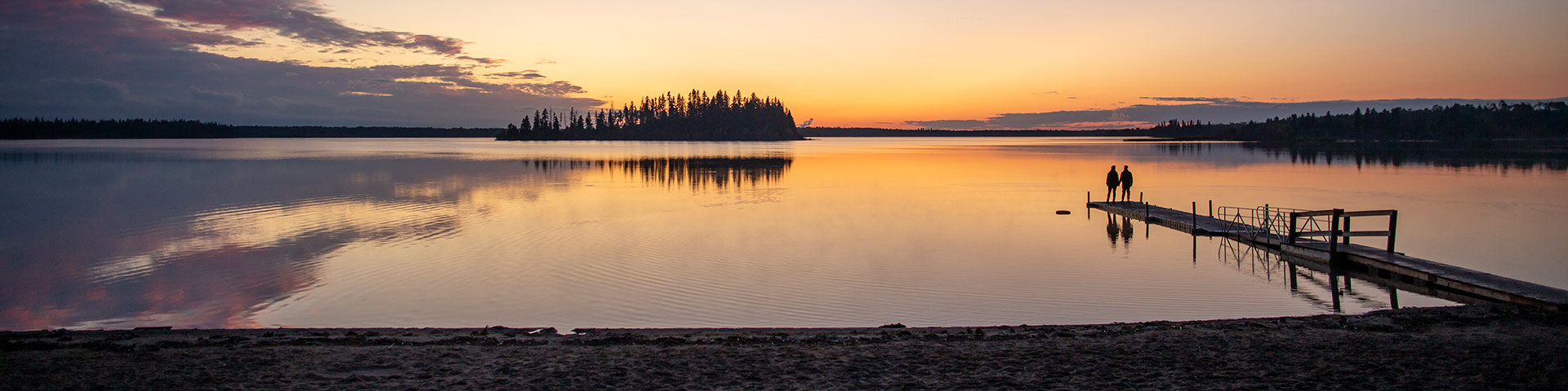 Two visitors stand on the dock and watch the sunset over Astotin Lake, Elk Island National Park.