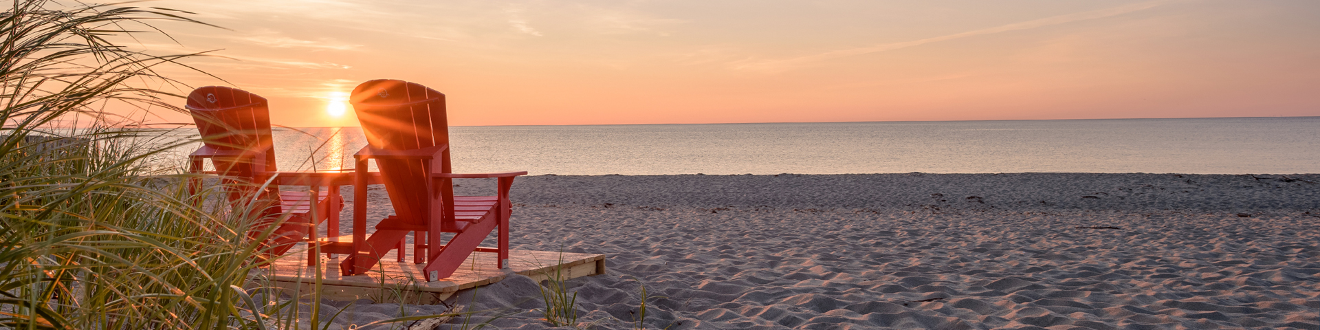 Two Parks Canada red chairs on a sandy beach at sunrise.