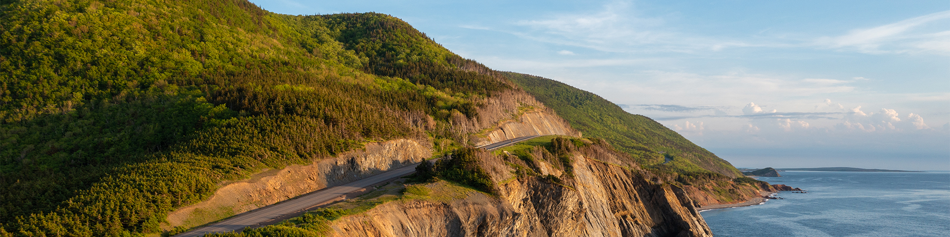 An aerial view of a cliff by the ocean with rolling green mountains in the background on the Cabot Trail.