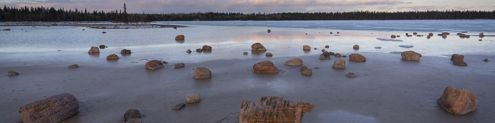Une photo du lac Grosbeak et des blocs erratiques (rochers) qui parsèment le sol mouillé. C’est le crépuscule et le soleil couchant donne au ciel une teinte et rose et bleue.