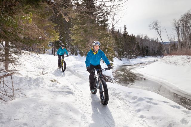 Deux amateurs de vélo à pneus surdimensionnés (fatbike) roulent sur un sentier de neige près d'une petite rivière.  