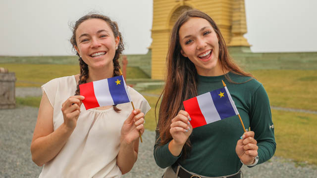 A uniformed Parks Canada guide smiles with a young woman infront of the yellow Frederic Gate at the fortress of Louisbourg. They are both holding small Acadian flags.