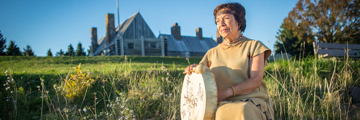 An indigenous woman in traditional Mi’kmaw clothing sits with a hand drum outside the Habitation at Port-Royal.