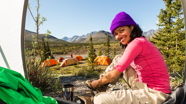 A woman in a tent looks at the camera, several tents and the mountains in the background at Ivvavik National Park.