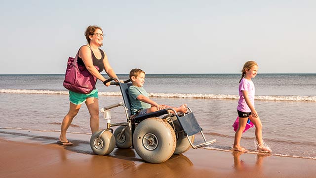 A beach wheelchair with large balloon tires, a sturdy metal frame, and a reclining seat is being pushed along a sandy shoreline
