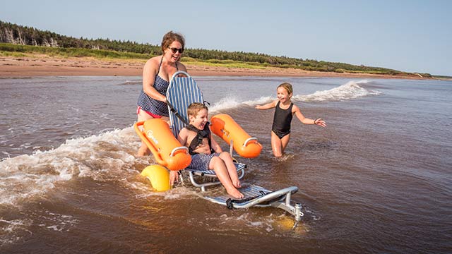 An excited child in a floating beach wheelchair is guided into the water by an adult, with another excited child nearby.