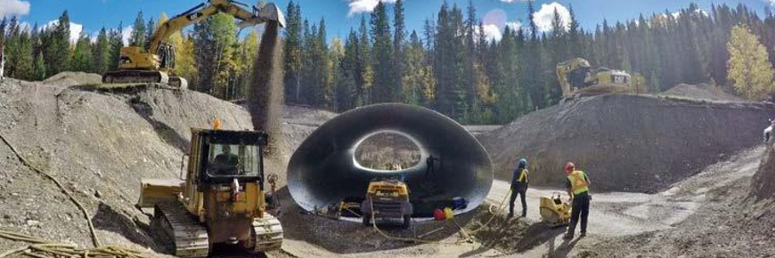 Des Travailleurs entourant une grande section de ponceau métallique et de l’équipement lourd.