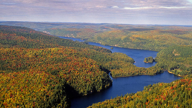 Vue aérienne d’un vaste paysage de collines boisées aux couleurs de l’automne entourant un lac bleu sinueux.