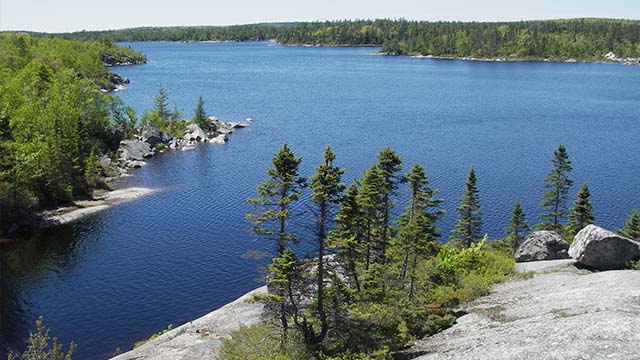 Un affleurement rocheux lisse, parsemé de conifères, fait saillie sur un lac calme au rivage rocheux, bordé d’une forêt boréale.