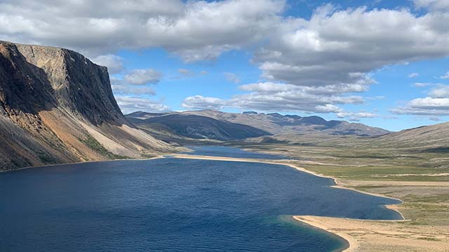 Une falaise abrupte descend dans un bras de mer calme, entouré de l’autre côté par des landes en pente. Au-delà s’étendent des montagnes ondulantes et un ciel de nuages.
