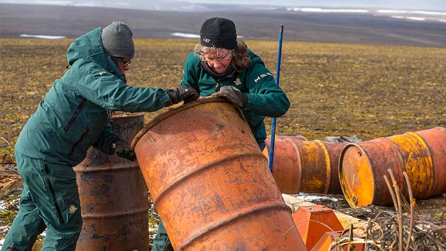 On a tundra landscape, Jane Chisholm and Angela Piercey stand up a large, rusty barrel, amid other discarded barrels and metal debris.