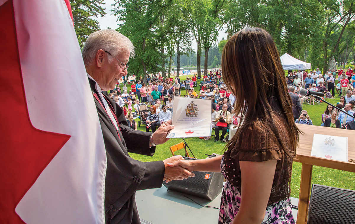Une femme reçoit son certificat de citoyenneté pendant une cérémonie extérieure au Parc national de Mont Riding. En arrière-plan, un public de visiteurs observe la scène.