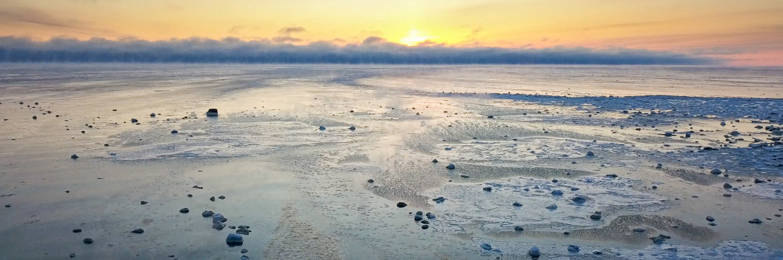 A view of the western James Bay landscape of frozen ice. The sun is setting in the background, with clouds starting to cover the yellow, orange, and pink sky.   