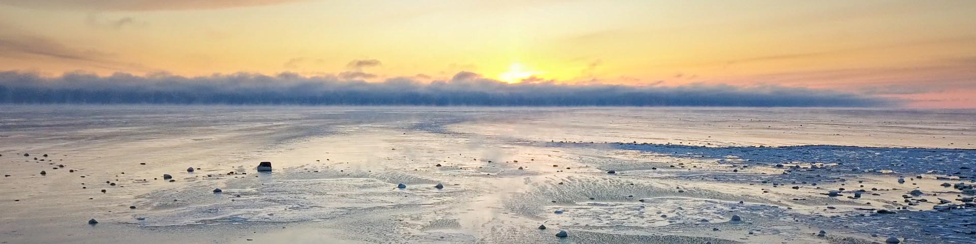 A view of the western James Bay landscape of frozen ice. The sun is setting in the background, with clouds starting to cover the yellow, orange, and pink sky.   