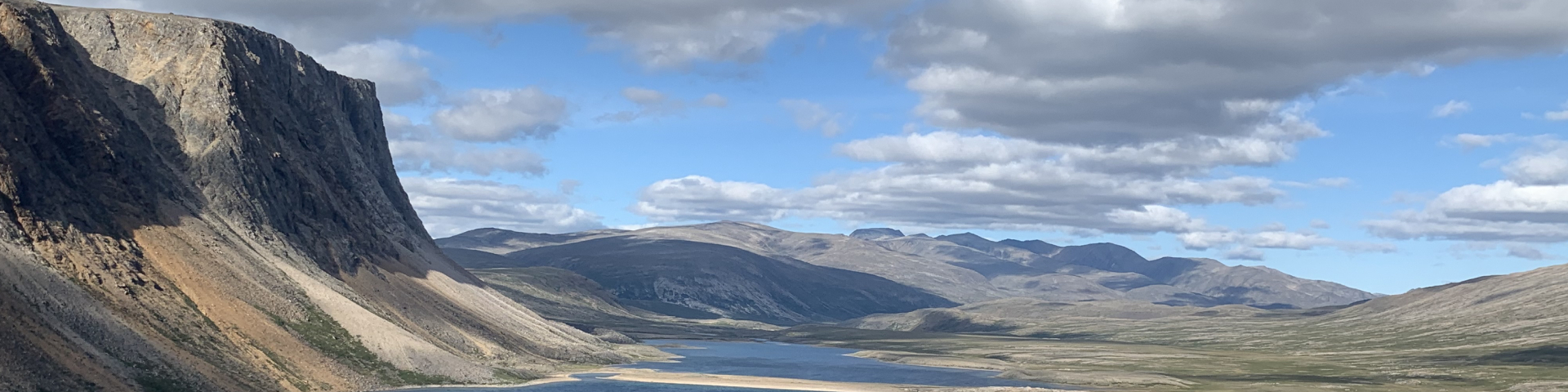 A scenic landscape featuring a large, deep blue lake surrounded by rugged mountains. The sky is bright blue with white clouds, and a narrow strip of land separates the lake from a larger body of water in the distance.