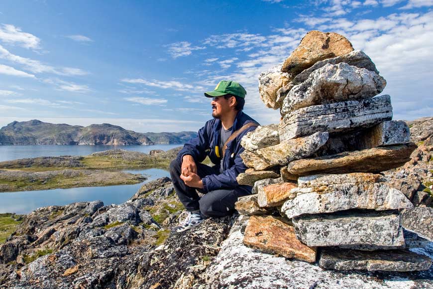 Indigenous man in a green ball hat sitting next to an inukshuk gazing at the adjacent mountains.