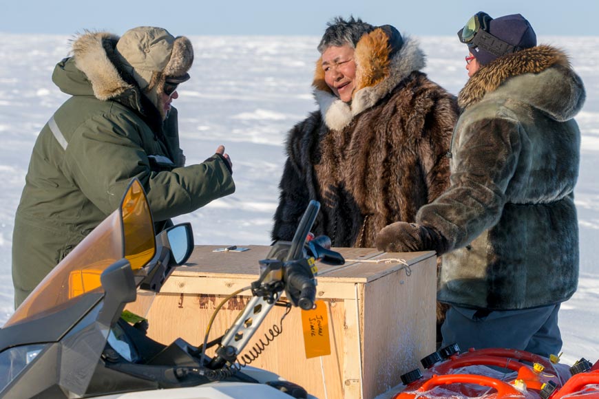Trois hommes autochtones portant des manteaux de fourrure, debout devant une machine à neige, dans la toundra arctique.