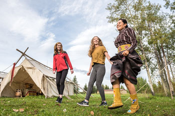 Two female visitors follow along to a Métis dance jig with an Indigenous woman leading the steps at the Métis Campfire station. Rocky Mountain House National Historic Site. 