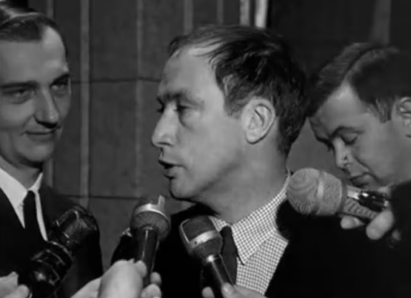 A man speaks into media microphone with two other men in the background