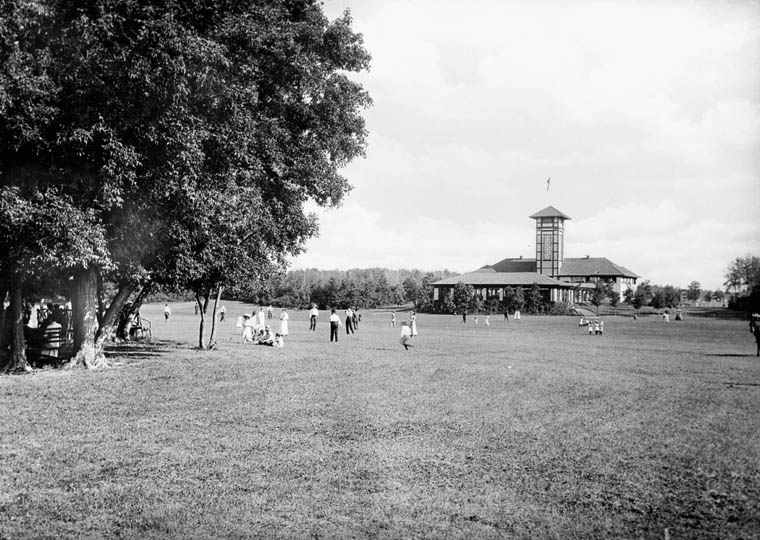 A photo of the Assiniboine Park Pavilion taken from a large grass field that children are playing on.