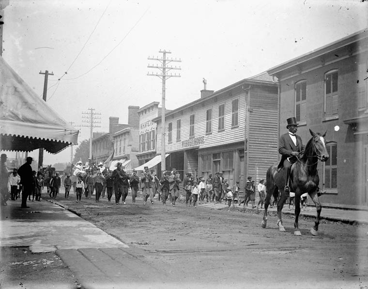Black and white photo of a parade in a street showcasing a person on a horse