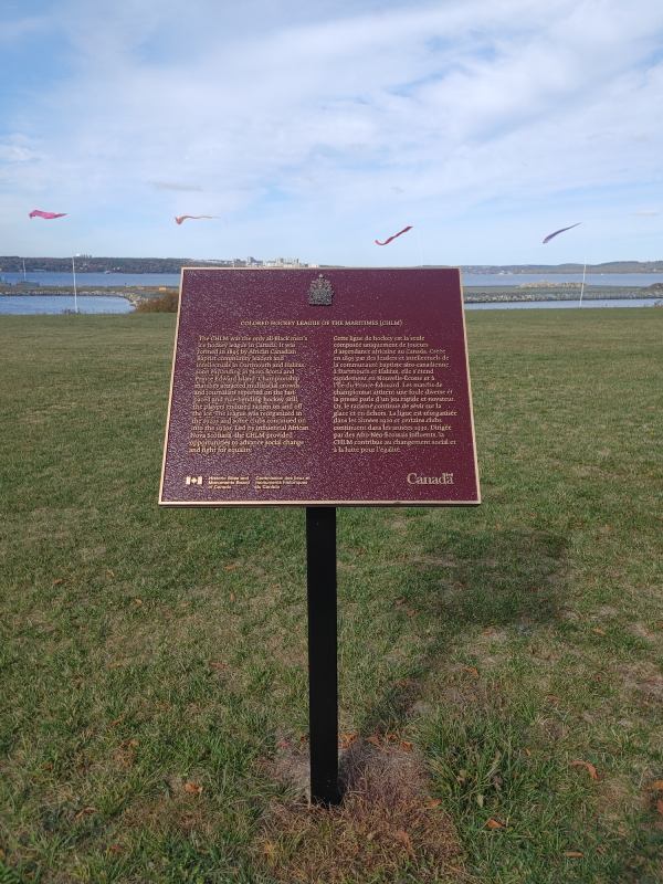 A commemorative plaque mounted on a black metal stand is installed on a grassy area overlooking a body of water