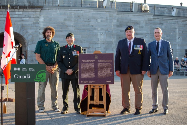 Four man, some wearing military uniforms, stand beside a commemorative plaque in from of a fort