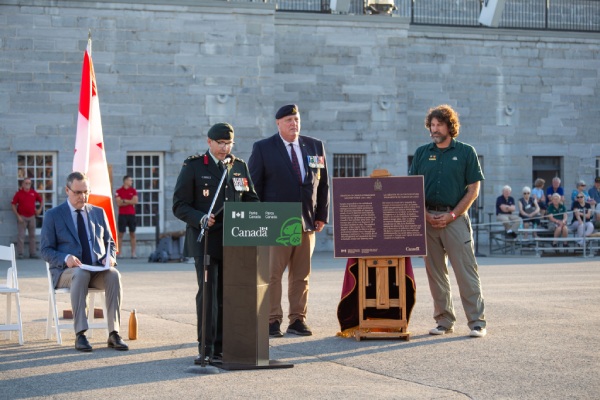 A man wearing a military uniform adresses a speach during an outdoor event in from of a fort, three other man behind him