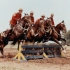 Four scarlet-uniformed police constables on horses riding over a fence