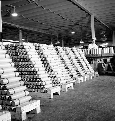 View of 25-pounder shells piled ready for shipment with workman at conveyor belt at Defence Industries Limited, 1944