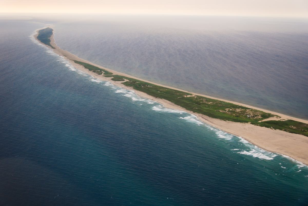 Aerial view of a narrow and long island made of sand a green grass surrounded by deep blue water