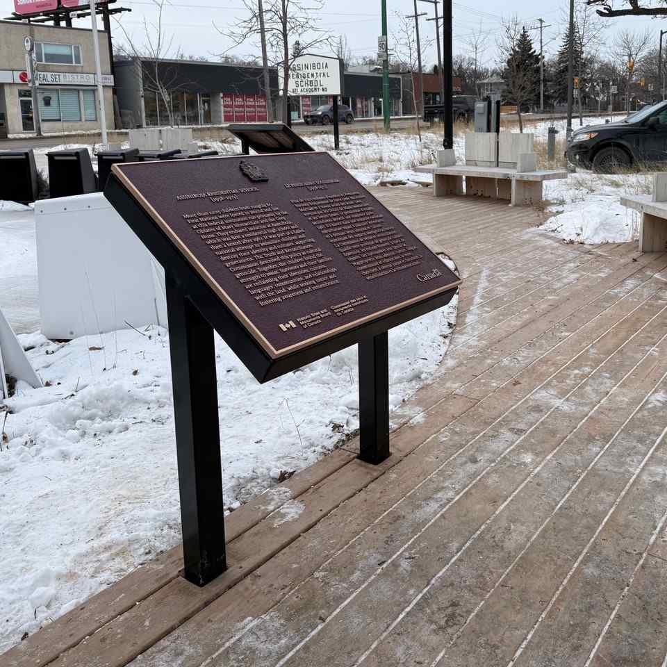 A commemorative plaque for Assiniboia Residential School (1958–1973) is installed in a wooden walkpath in a town
