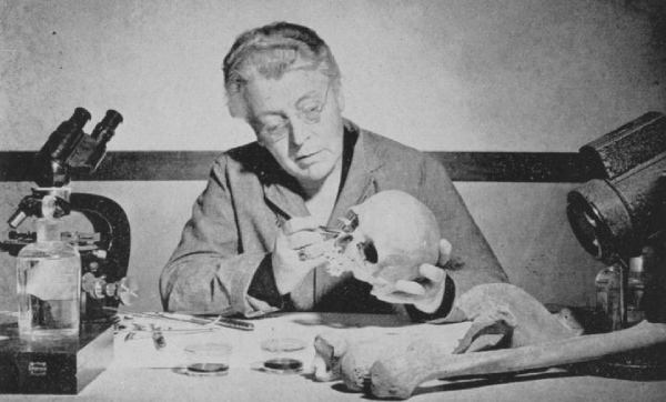 A woman handles bones, sitting at a desk