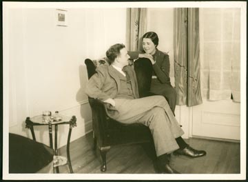 Un homme est assis, les jambes croisées, et regarde une femme assise sur le bras de sa chaise. 