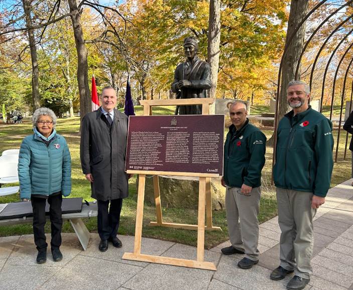 Four persons stand beside a commemorative plaque installed on an easel in a fall forest setting