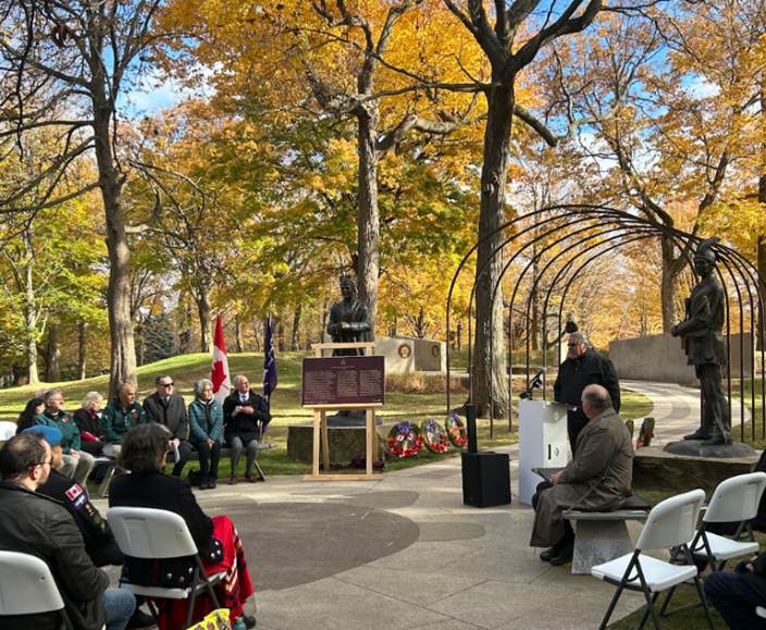 An outdoor autumnal ceremony where people are seated and watching someone speak to a lectern