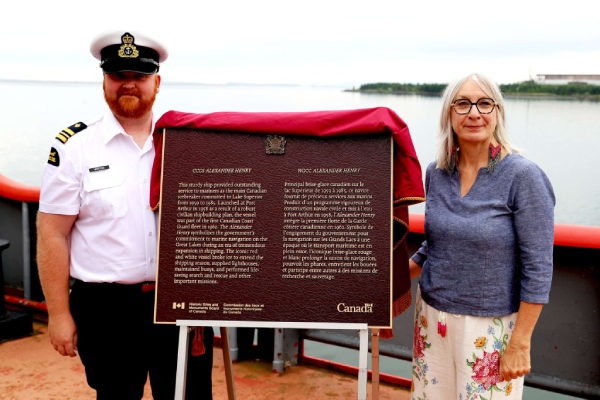 Two persons, one wearing a uniform, stand on the deck of a ship beside a commemorative plaque