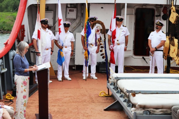 Uniformed man on the deck of a ship stand as a women speaks at a lectern