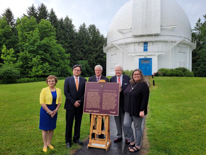 A group of people beside a commemorative plaque, in front of a astronomy observatory