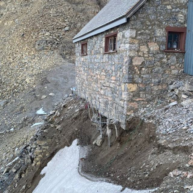 The ground beneath the north facade of a stone cabin is eroding, leaving empty spaces beneath its foundations.
