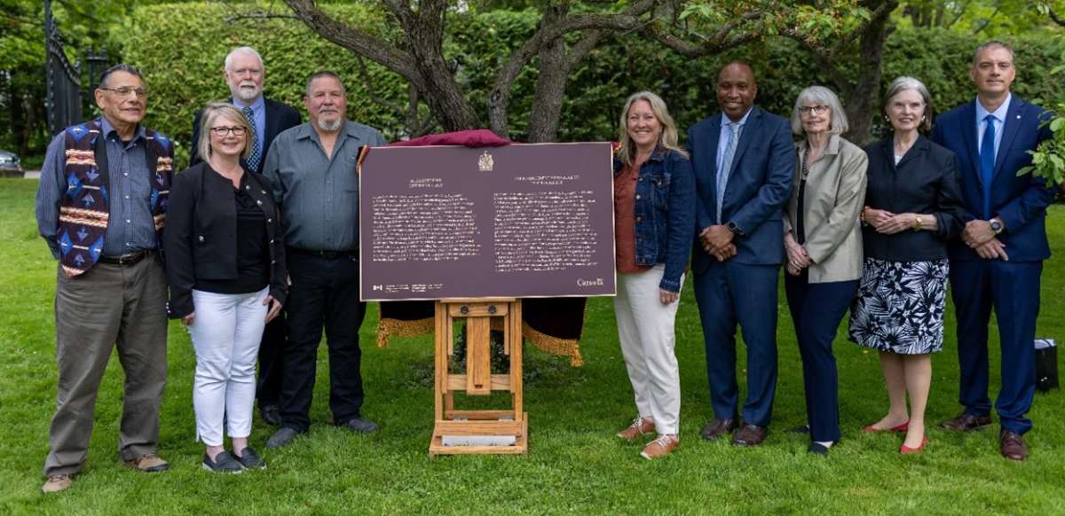 A group of people stand near a commemorative plaque, against a backdrop of summer greenery