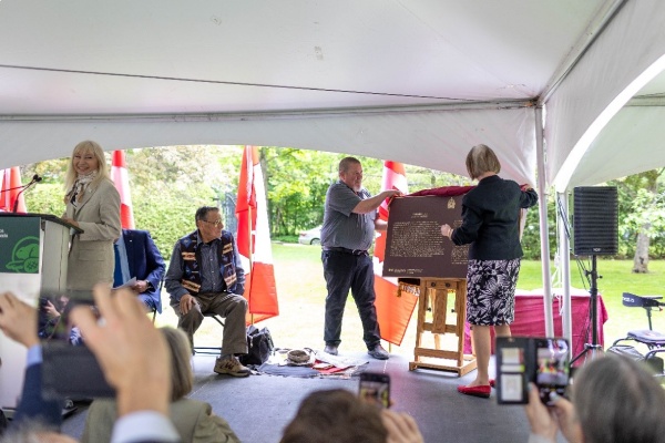 Two people unveil a commemorative plaque in front of a crowd under a marquee