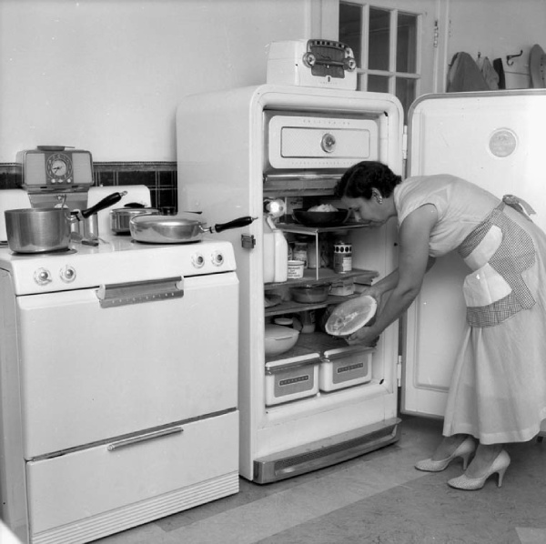 A woman in the 1950s opens a refrigerator in a kitchen