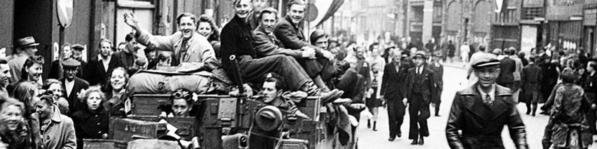 Black-and-white photograph of civilians riding on a military tank during a wartime liberation celebration, surrounded by crowds in a city street.