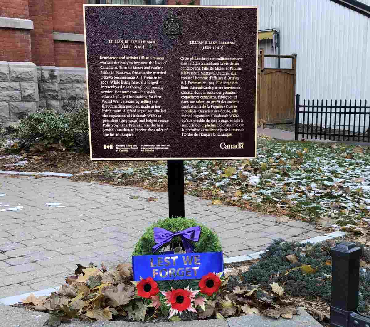 Plaque commémorative fixée sur un poteau noir devant un bâtiment, avec des fleurs déposées à sa base