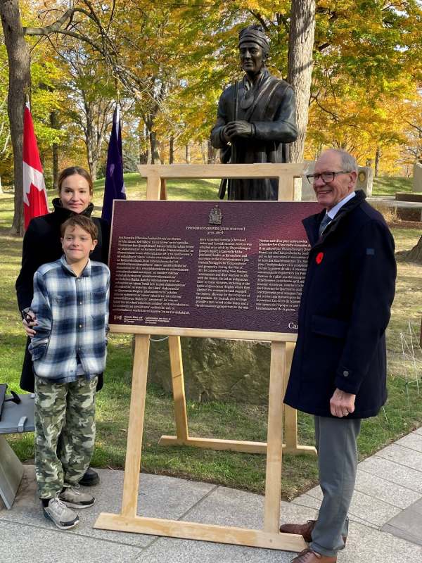 Deux adultes et un enfant se tiennent à côté d'une plaque commémorative dans un environnement extérieur automnal